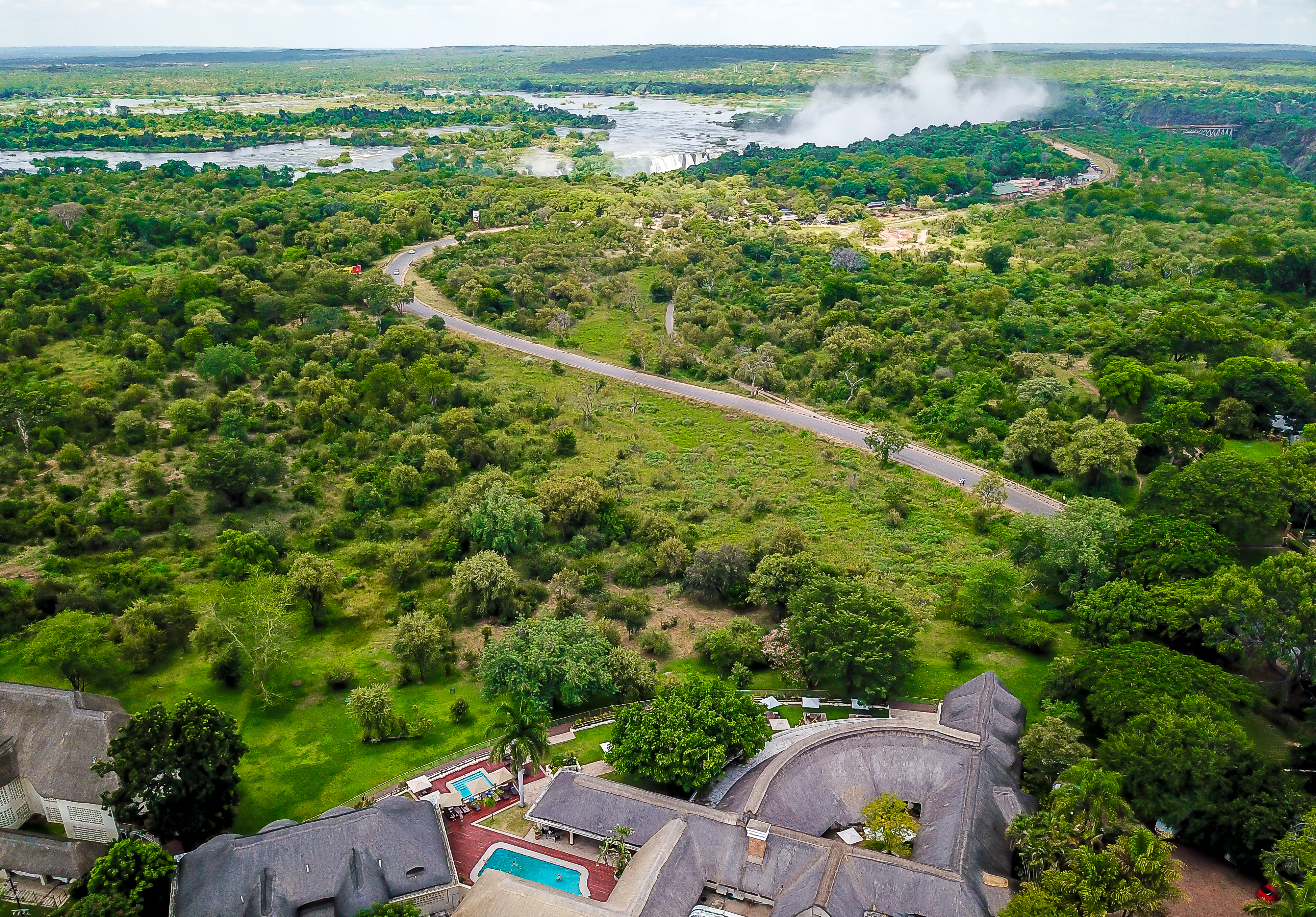 Aerial view over the Zambezi River, lush rainforest and Victoria Falls spray with a safari lodge and pool in the foreground
